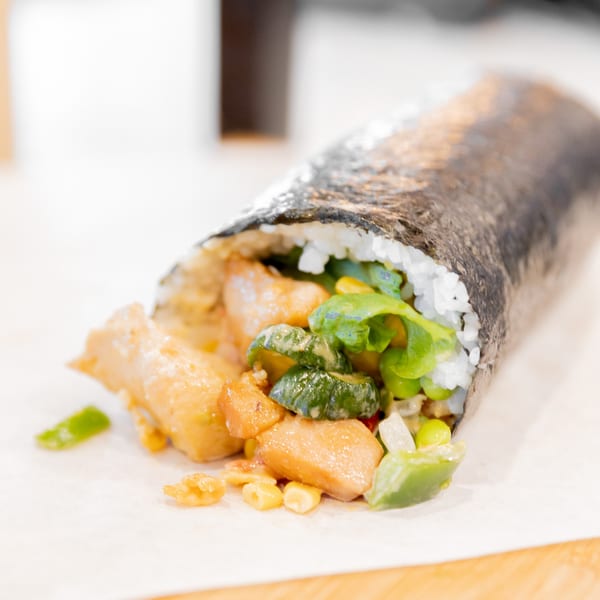 Nooki sushi burrito with chicken, zucchini, corn, and rice spilling out of the nori wrap.